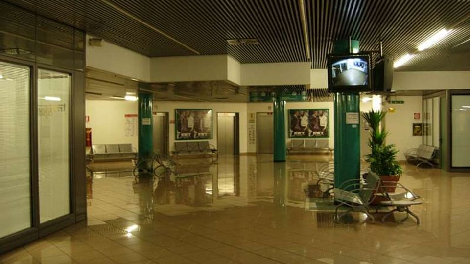 Ampia hall interna con pavimento lucido in piastrelle, soffitto a listelli scuri e pilastri verdi, con panche metalliche e alcune piante in vaso. Sulle pareti sono presenti poster e, in alto, un monitor di sorveglianza; sul fondo si vedono porte e corridoi di accesso.