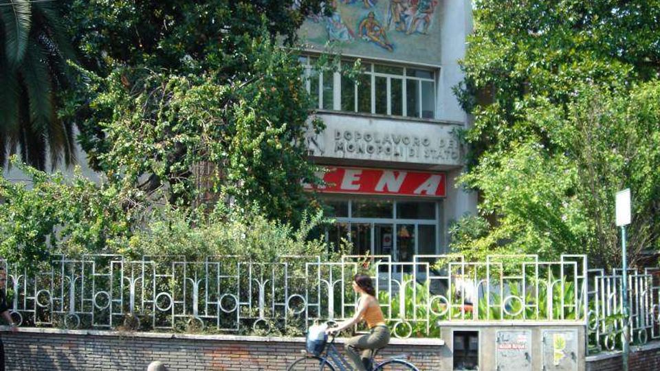 Facciata di un edificio pubblico parzialmente nascosta da alberi e vegetazione, con grande finestra centrale e un murale decorativo nella parte alta. Davanti si vede una recinzione metallica sopra un muretto in mattoni lungo il marciapiede e la strada.