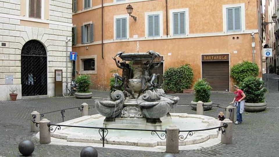 Piccola piazza lastricata con una fontana monumentale in pietra al centro, con vasca ottagonale e sculture decorative. Sullo sfondo si vedono edifici storici intonacati con persiane, un portone ad arco e vasi con arbusti lungo i bordi della piazza.
