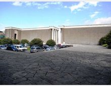 Grande edificio moderno in cemento con facciata lunga e minimale, interrotta da un portale e da alte colonne verticali. In primo piano si vede una piazza lastricata in pietra con parcheggio di auto e alberi ai margini sotto un cielo parzialmente nuvoloso.