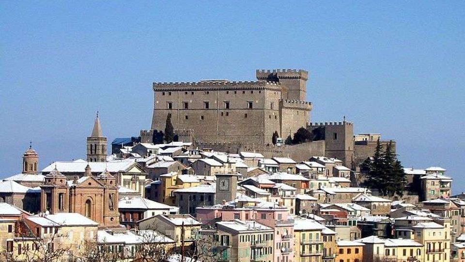 Veduta panoramica di un borgo collinare con case compatte dai tetti innevati e una grande rocca in pietra merlata in cima. In primo piano si distinguono edifici colorati e una chiesa con campanile, sullo sfondo cielo limpido.