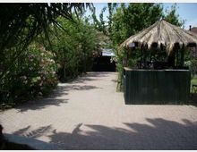Vialetto pavimentato in un’area verde con alberi e cespugli fioriti ai lati, che conduce verso una struttura sullo sfondo. A destra si vede un chiosco con tetto in paglia e bancone in legno scuro, in un contesto tipo giardino o cortile.