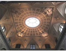 Soffitto interno di un edificio storico con grande lucernario ovale centrale, circondato da cassettoni decorati e rilievi scultorei agli angoli. Lungo il perimetro si vedono archi a sesto acuto e finestre superiori incorniciate da stucchi ornamentali.