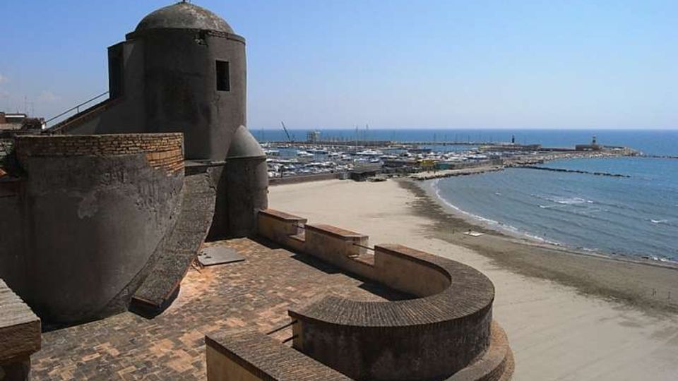 Veduta costiera con una fortificazione in muratura e torre semicircolare in primo piano, affacciata su una lunga spiaggia sabbiosa. Sullo sfondo si vedono il mare, un porto con numerose barche e un lungo molo frangiflutti.