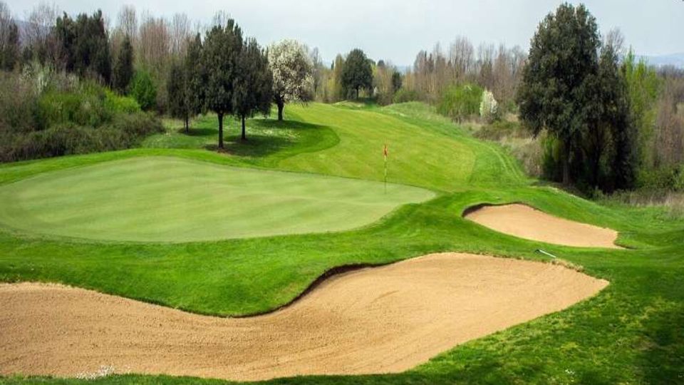 Campo da golf con ampio green e bandierina, circondato da fairway ondulato e alberi sullo sfondo. In primo piano si vedono due bunker di sabbia con bordi curvi lungo il lato destro del green.