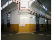 Interno di un capannone industriale con soffitto alto e travi a volta, grandi finestre ad arco e porte metalliche blu lungo le pareti. Pareti bianche e ocra con tubazioni a vista rosse e grigie vicino al soffitto e pavimento in cemento segnato.