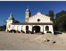 Chiesa bianca a un piano con portico ad archi, rosone centrale e facciata con profilo ondulato, affiancata da una torre campanaria con orologio e cupola verde. Davanti si estende un ampio piazzale in ghiaia con vasi di piante, sullo sfondo alberi e cielo sereno.