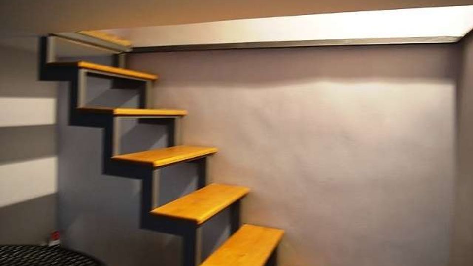 Scala interna compatta con gradini in legno a sbalzo, fissati a una struttura metallica contro una parete grigia. L’ambiente ha soffitto basso e illuminazione dall’alto, con pavimento in piastrelle scure.