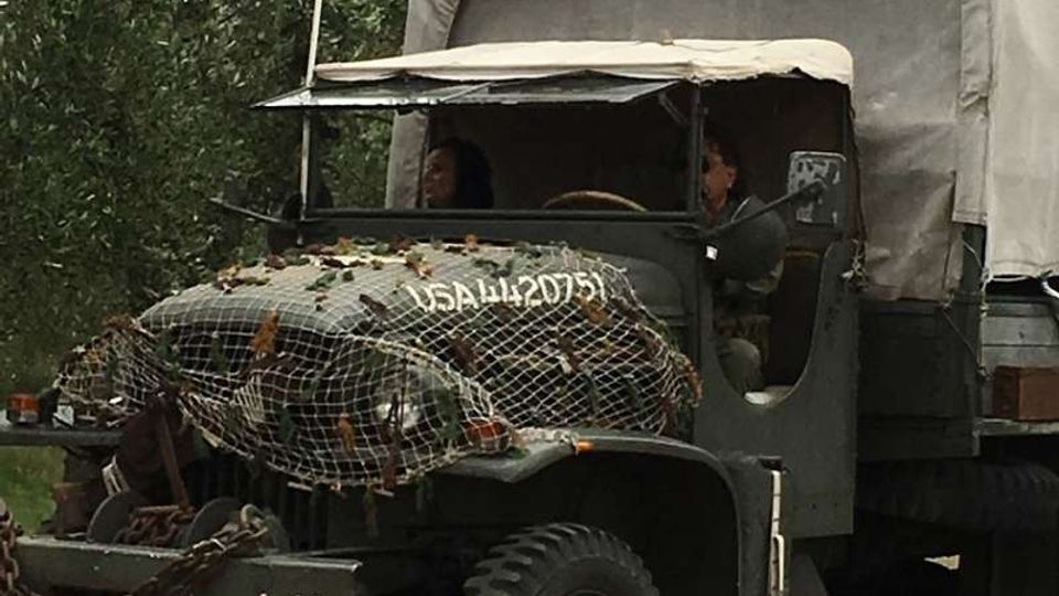 Camion militare d’epoca con telone sul cassone e rete mimetica sul cofano, fermo su una strada sterrata circondata da alberi. Una bandiera statunitense è montata su un’asta nella parte anteriore del veicolo.