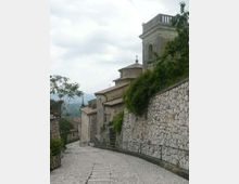 Strada in pendenza lastricata in pietra in un borgo collinare, affiancata da un alto muro in pietra e vegetazione. Sul lato destro si vede una chiesa in muratura con tetti in coppi e un campanile, con montagne sullo sfondo.