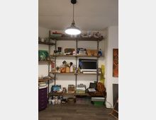 Angolo di studio/laboratorio artistico con pareti bianche e scaffalature in legno e metallo piene di barattoli, colori, pennelli e materiali da lavoro. Una lampada a sospensione illumina un ripiano con un monitor e, sotto, contenitori e scatole organizzate su pavimento in legno.