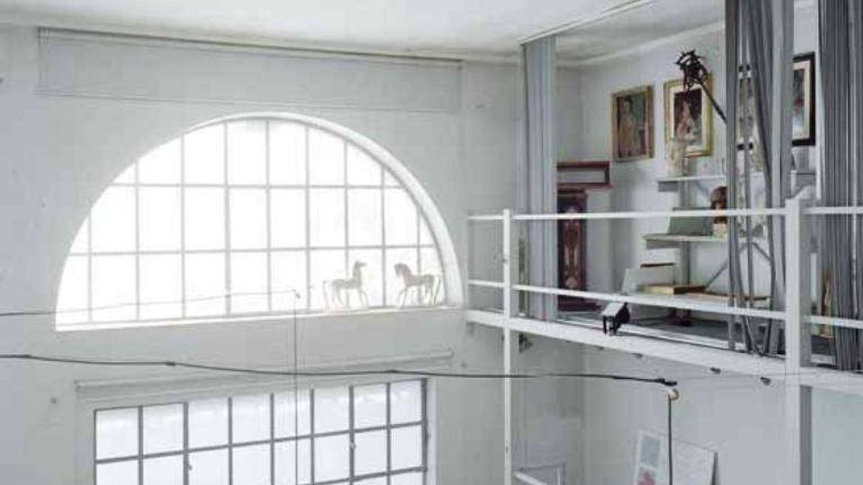 Interno di un loft a doppia altezza con soppalco e ringhiere bianche, grandi finestre a griglia e una finestra ad arco che illuminano lo spazio. Sul soppalco si vedono opere e cornici appoggiate e pareti con quadri, in un ambiente in stile industriale.