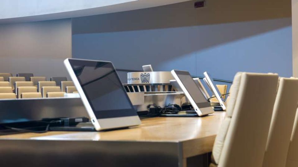Sala conferenze moderna con file di sedute sullo sfondo e un grande tavolo in legno in primo piano con schermi/monitor inclinati e cablaggi. Pareti chiare e soffitto curvo con illuminazione integrata definiscono un ambiente istituzionale e ordinato.