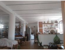 Interno open space con cucina a vista e bancone in muratura rivestito di piastrelle, illuminato da finestre laterali e lampade a sospensione. Soffitto con travi dipinte di bianco, colonne, pavimento in piastrelle e arredi misti tra tavoli, sedie e scaffali con oggetti decorativi.