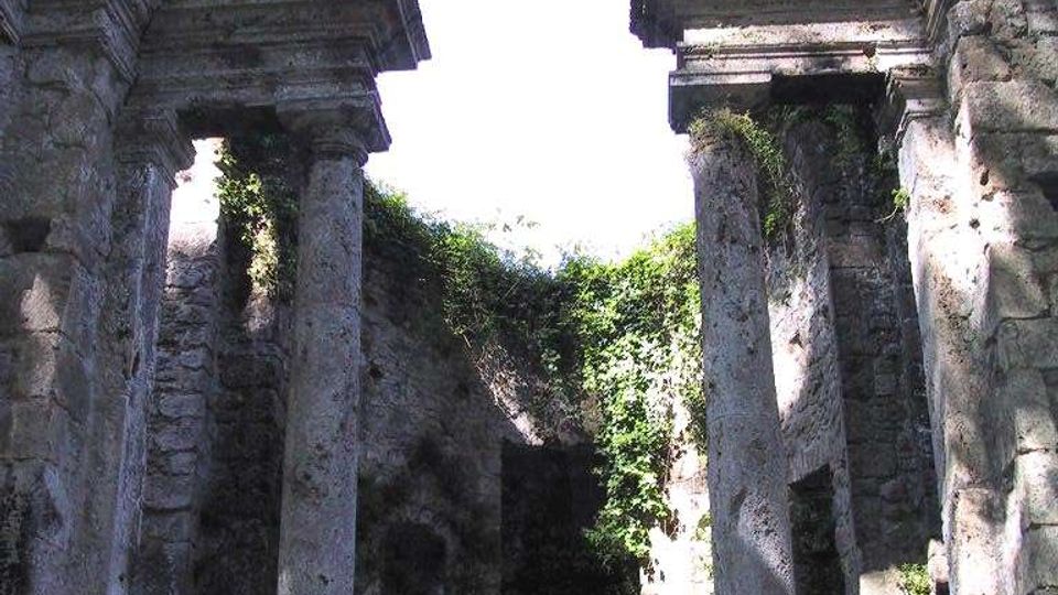 Interno in rovina di un edificio storico in pietra, con grande arco semicircolare decorato da oculi e due alte colonne con capitelli. La struttura è a cielo aperto, con pareti parzialmente crollate e vegetazione che cresce tra le murature.