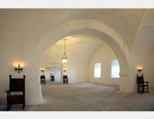 Ampia sala con soffitto a volte bianche e grandi archi in muratura, pavimento in pietra e finestre rettangolari lungo la parete destra. L’ambiente è illuminato da un lampadario centrale e da applique a parete, con alcune sedie in legno disposte lungo i lati.