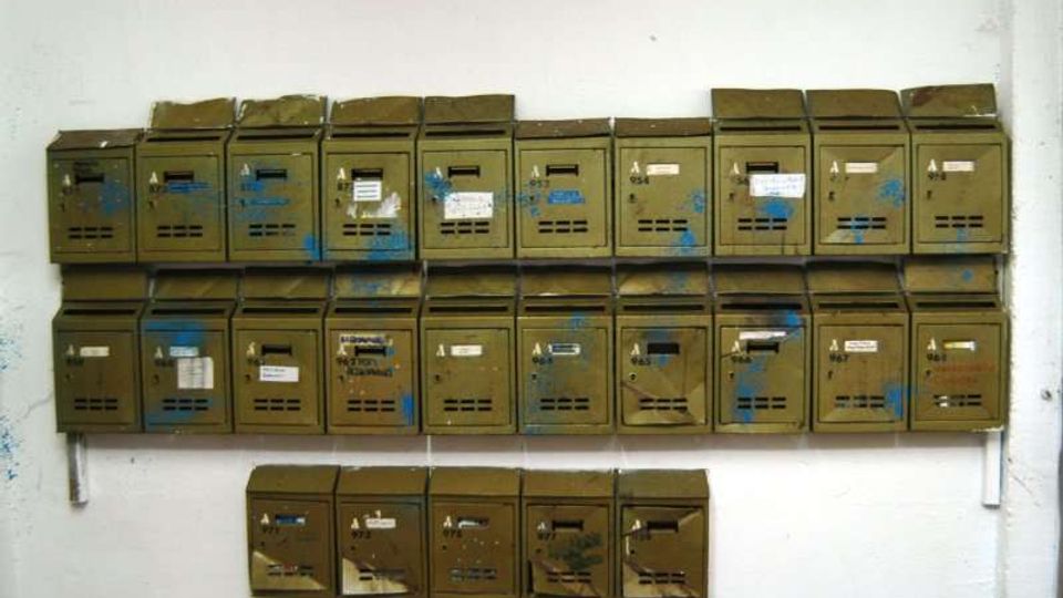 Fila di cassette postali metalliche verde oliva fissate a una parete bianca, disposte su tre livelli; molte hanno etichette, fessure per la posta e segni di usura o vernice.