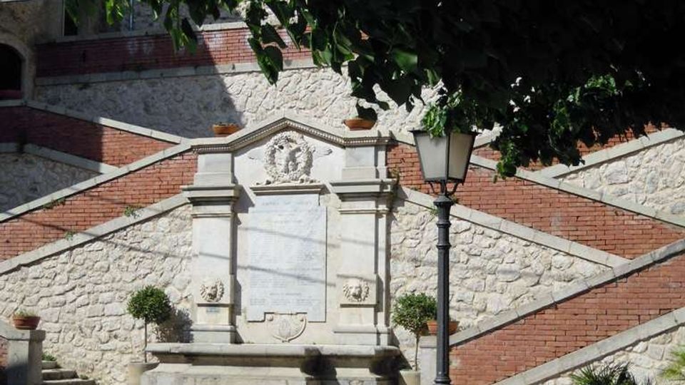 Monumento in pietra con lapide incassata in un muro a terrazze rivestito di pietra e fasce di mattoni, affiancato da una scalinata. In primo piano si vedono un lampione in ferro, muretti e vasi con piante sotto la chioma di alberi.