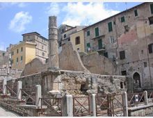 Rovine archeologiche in pietra con una colonna antica isolata e muri parzialmente crollati, circondate da una recinzione metallica. Sullo sfondo si vedono edifici storici in muratura con persiane verdi affacciati sulla piazza sotto un cielo parzialmente nuvoloso.