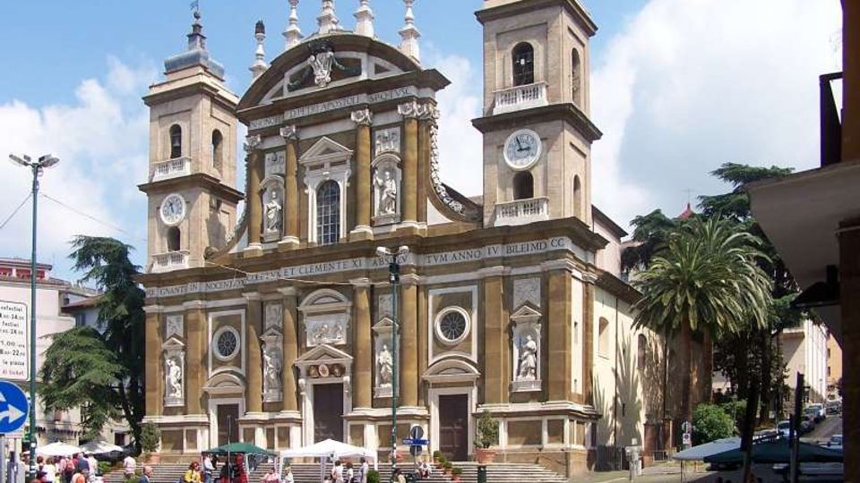 Facciata di una grande chiesa in stile barocco con due campanili laterali dotati di orologi, statue in nicchie e frontone decorato con croci e pinnacoli. Davanti si apre una piazza con gradinate, bancarelle e alberi, circondata da edifici urbani.