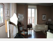 Soggiorno arredato con divani chiari, poltrona e tavolino su pavimento in legno scuro, con quadri alle pareti e lampade da tavolo. Al centro una portafinestra con tende apre su un balcone con vista su edifici esterni.