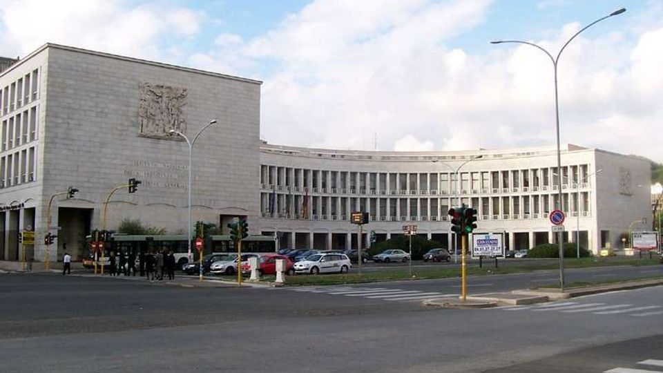 Grande edificio istituzionale moderno in pietra chiara, con un corpo principale rettangolare e un’ala curva a più piani con file regolari di finestre. In primo piano si vede un ampio incrocio urbano con semafori, strisce pedonali, auto parcheggiate e lampioni sotto un cielo parzialmente nuvoloso.