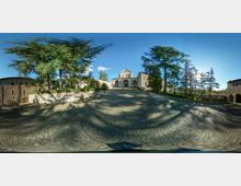 Ampia piazza in ciottoli in pendenza che conduce a un complesso in pietra con portico ad archi e facciata semplice, affiancato da edifici storici. Grandi alberi e giardini incorniciano lo spazio sotto un cielo sereno.
