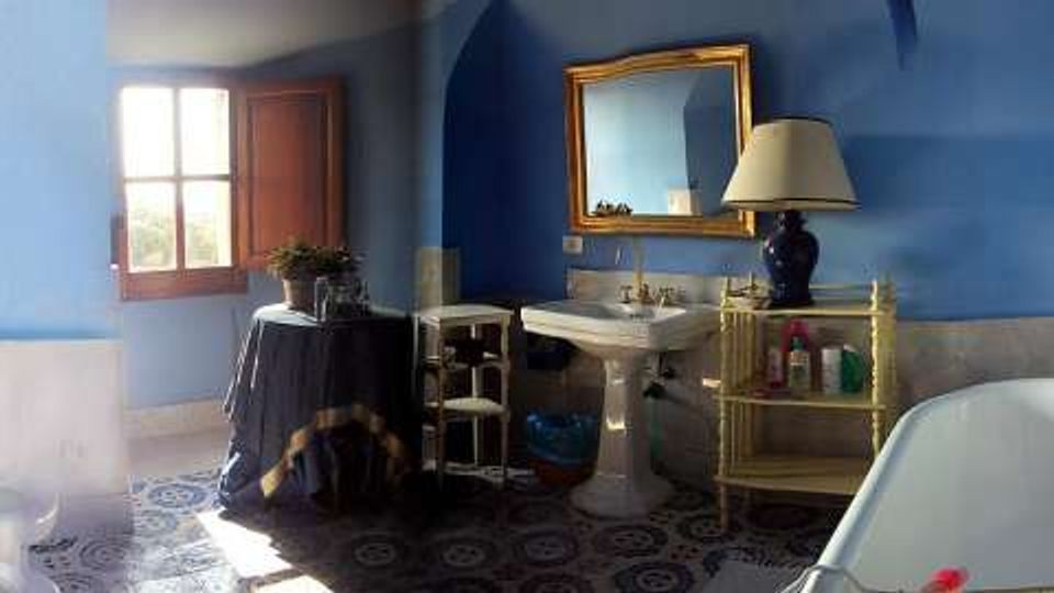 Bagno con pareti blu e pavimento in piastrelle decorative, dotato di vasca con rubinetteria in ottone, lavandino su colonna e grande specchio con cornice dorata. Una finestra in legno lascia entrare luce naturale e accanto al lavandino ci sono un mobiletto e una lampada da tavolo.