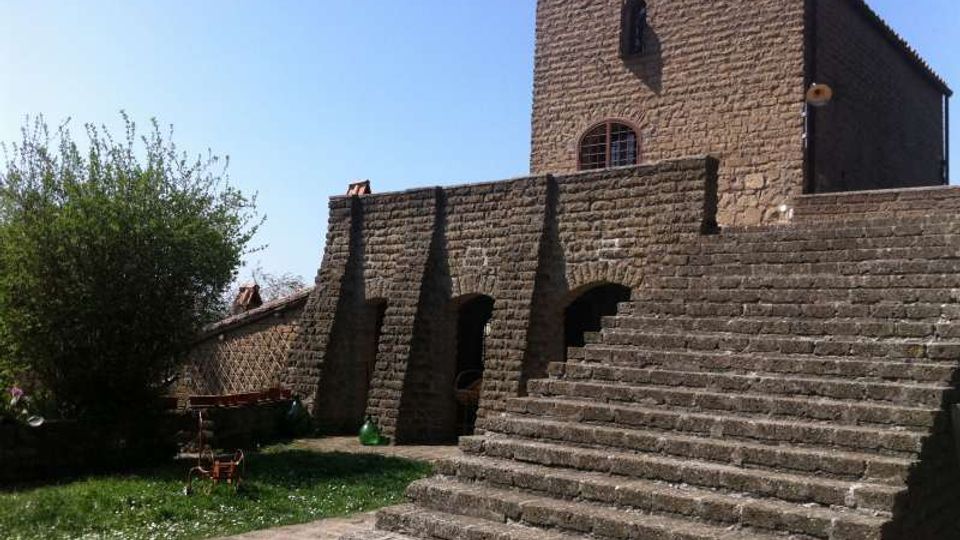Edificio in pietra con facciata semplice e una finestra ad arco, affiancato da un’ampia scalinata in pietra e da arcate alla base. Intorno si vede un piccolo prato con alberi e un cortile esterno sotto cielo sereno.