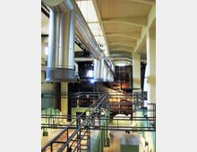 Interno di un grande edificio industriale riconvertito, con soffitto alto a travi e grandi condotti metallici di ventilazione sospesi. Passerelle e ringhiere metalliche attraversano lo spazio sopra un’area espositiva con pareti verdi e sculture su piedistalli.