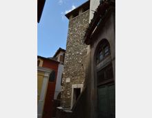 Torre alta in pietra grezza con piccole aperture in alto, affacciata su un vicolo stretto tra edifici colorati. A destra si vede una facciata con grande portone ad arco e balcone con fioriere sotto il tetto.