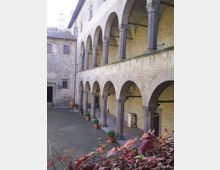 Cortile interno di un edificio storico in pietra con portico a due livelli, caratterizzato da file di archi in mattoni sostenuti da colonne grigie. Lungo il porticato sono disposti vasi in terracotta con piante, con muri in pietra e tetti in coppi sullo sfondo.