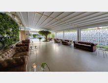 Ampia lounge moderna con soffitto a pannelli e grandi vetrate su un lato, schermate da pannelli decorativi bianchi. Divani in pelle scura con tavolini in metallo e pareti verdi con piante creano un’area relax luminosa.