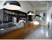 Interno di un bar moderno con lungo bancone in legno e vetrina, parete nera con scaffali di bottiglie e grandi lampade a sospensione a cupola. Pavimento chiaro lucido, soffitto alto e grandi finestre sul fondo che illuminano l’ambiente.