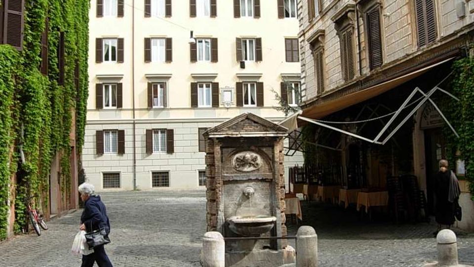 Piccola piazza acciottolata tra edifici storici con persiane e facciate chiare, con una fontana in pietra al centro. A sinistra una parete ricoperta di edera e a destra un portico con tende davanti a un locale.