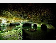 Interno di una grotta carsica con soffitto basso e irregolare e pareti rocciose coperte di muschio. Un corso d’acqua scorre sul fondo formando pozze scure, con aperture ad arco nella roccia illuminate da luce artificiale.