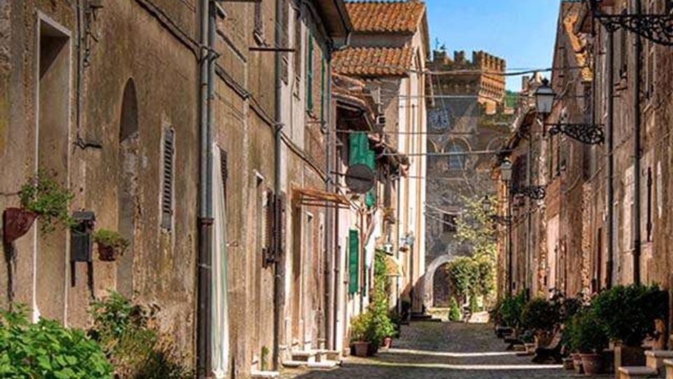Stradina stretta acciottolata in un borgo storico, fiancheggiata da case in pietra con intonaco consumato, persiane e balconcini. Lungo i lati ci sono vasi di piante, lampioni a parete e, sullo sfondo, un arco e un edificio con tetto in tegole sotto un cielo sereno.