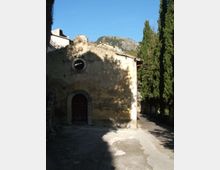 Facciata in pietra di una piccola chiesa o cappella con portale ad arco e finestra circolare, affacciata su un cortile in ombra. A destra corre un vialetto alberato con cipressi, con colline rocciose sullo sfondo sotto cielo sereno.