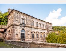 Edificio storico in pietra e intonaco, a due piani, con facciata decorata da cornici e nicchie e una grande porta ad arco con vetrate. In primo piano si vede un parapetto in pietra e un lampione a più bracci, con alberi e colline boscose sullo sfondo.