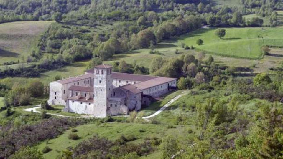 Veduta panoramica di un monastero in pietra con tetti in laterizio e torre quadrata centrale, situato in una valle. Intorno si vedono prati verdi, campi coltivati e colline boscose attraversate da una strada sterrata.