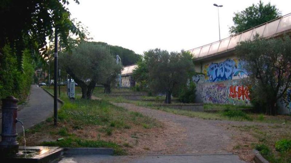 Percorso pedonale sterrato tra alberi e vegetazione, affiancato a destra da un lungo edificio con pareti coperte di graffiti e tetto inclinato. A sinistra si vede un marciapiede con lampioni e una fontanella su una piccola area pavimentata.