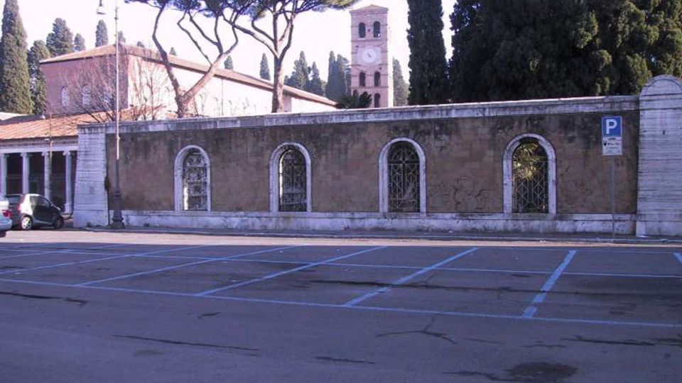 Parcheggio su strada con stalli segnati in blu davanti a un lungo muro in pietra con finestre ad arco e grate. Sullo sfondo si vedono alti cipressi, pini a ombrello e un campanile in mattoni con orologio.