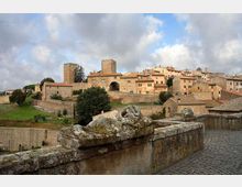 Vista panoramica di un borgo medievale su una collina, con case in pietra dai tetti in tegole, mura fortificate e una torre quadrata. In primo piano si vede un parapetto in pietra e un camminamento lastricato, con campi verdi ai piedi delle mura sotto un cielo nuvoloso.