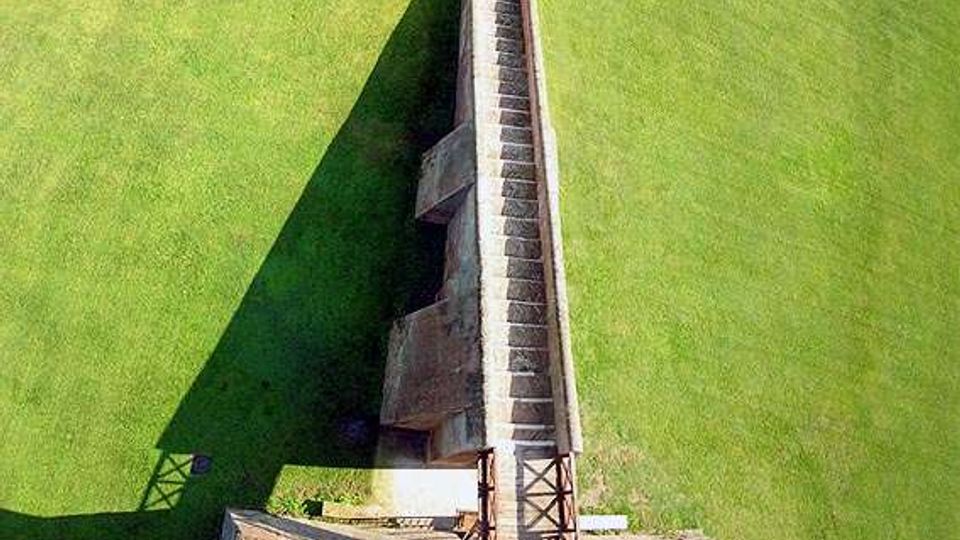 Veduta dall’alto di una lunga struttura in cemento a forma di rampa con gradini e pareti laterali, collocata al centro di un ampio prato verde. Alla base si vede una piattaforma con elementi metallici e un bordo in cemento che delimita l’area.
