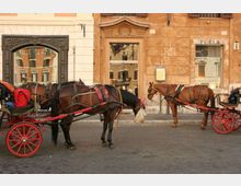 Strada urbana con pavimentazione in pietra davanti a una facciata di edifici storici con grandi portali e finestre. In primo piano ci sono carrozze trainate da cavalli con ruote rosse parcheggiate lungo il bordo della strada.