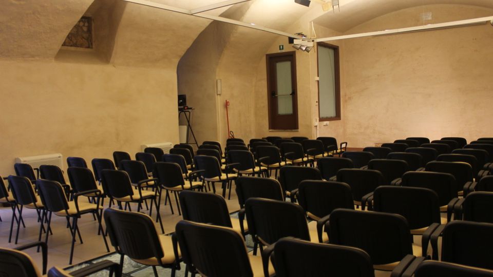 Sala conferenze interna con soffitto a volta e pareti color crema, illuminata da faretti su binari. File di sedie nere disposte in platea di fronte a una parete, con finestre e una porta sul lato.