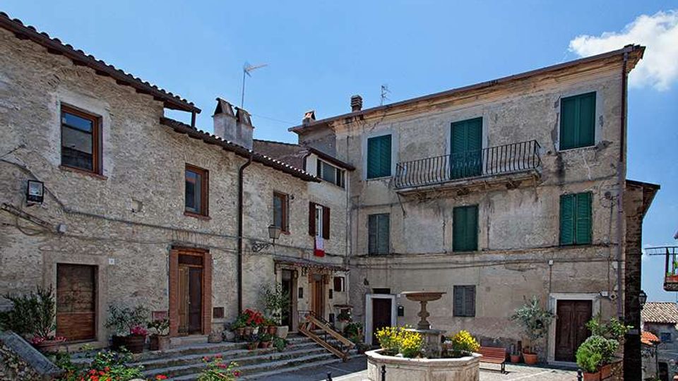 Piccola piazza in un borgo con case in pietra e intonaco, finestre con persiane verdi e balcone in ferro. Al centro c’è una fontana circolare in pietra, con gradini, panchine e fioriere, sotto un cielo sereno.
