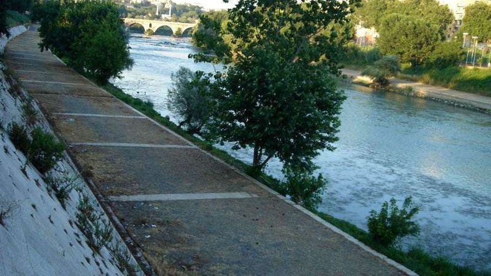 Passeggiata lungo un fiume con ampio argine in cemento e sentiero in ghiaia sul lato sinistro, affiancato da alberi e vegetazione. In lontananza si vede un ponte ad archi che attraversa il fiume, con la città sullo sfondo.