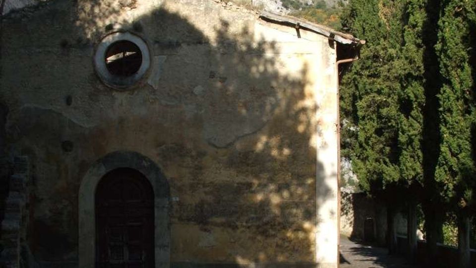 Facciata in pietra di una piccola chiesa o cappella con portale ad arco e finestra circolare, affacciata su un cortile in ombra. A destra corre un vialetto alberato con cipressi, con colline rocciose sullo sfondo sotto cielo sereno.