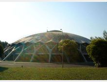 Grande edificio a cupola tipo palazzetto dello sport, con struttura esterna a travi triangolari e ampie vetrate alla base. Davanti si vedono una strada, un lampione e un’area verde con alberi ai lati.